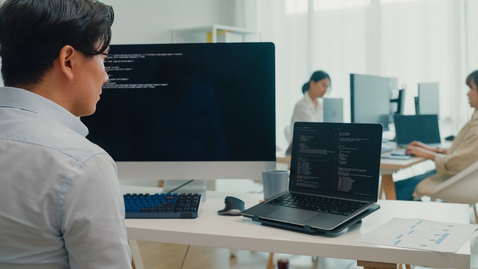Software developer working on dual screens with coworkers at desks in a bright office setting.