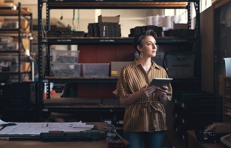 Woman in a striped shirt holding a tablet while standing thoughtfully in a well-organized warehouse.
