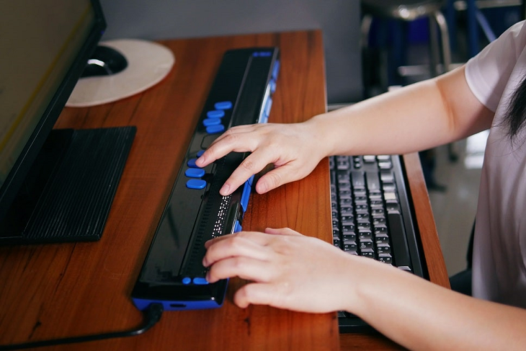 Person using a braille display with a computer keyboard on a wooden desk