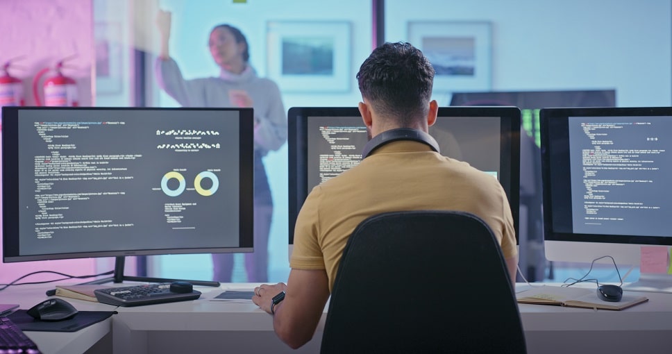 Developer working on system and API integration with multiple monitors displaying code, in a tech office environment.