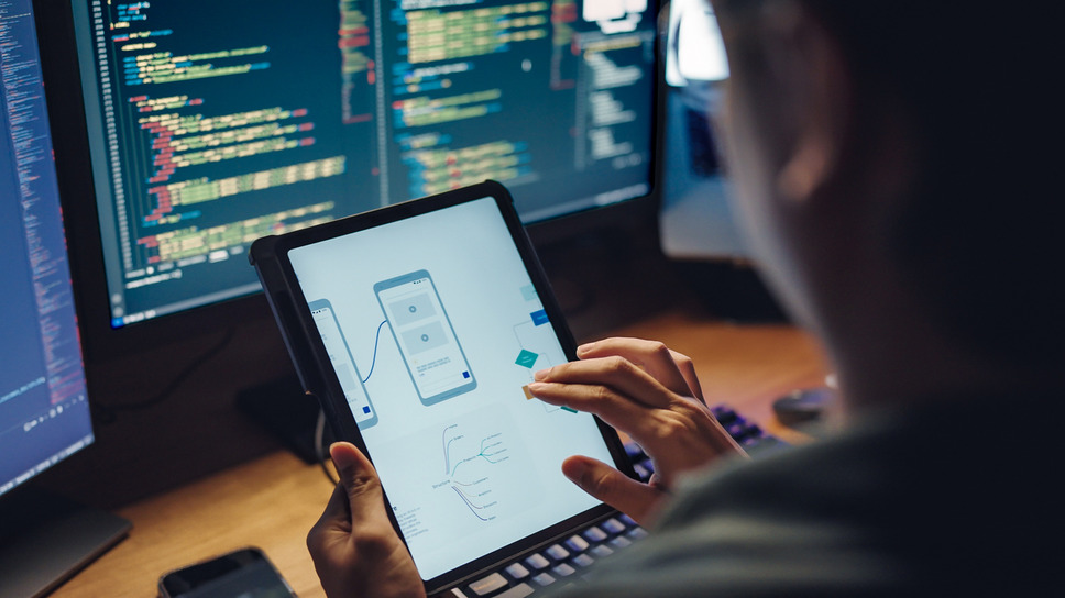 Developer analyzing mobile app interface on a tablet with code screens in background, illustrating app development process