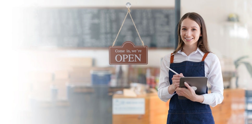 Smiling woman with a tablet in a cafe with an "Open" sign in the background.