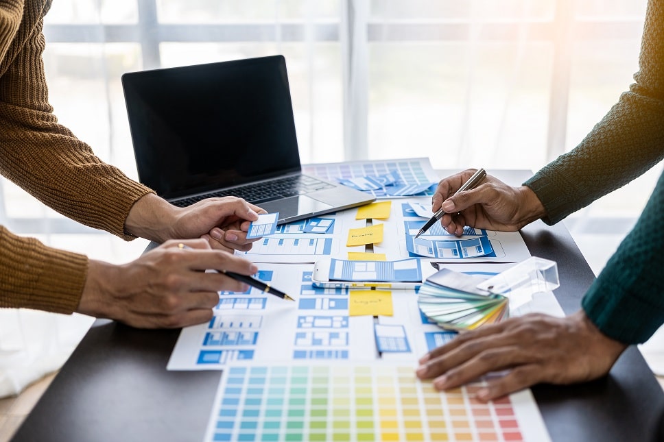 Two professionals collaborating on a web design project with color samples, website wireframes, and a laptop on the table