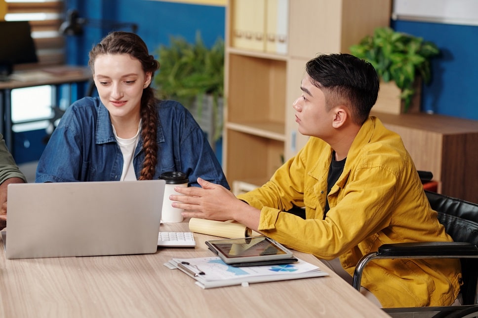 Two individuals in a collaborative work setting, one pointing at a laptop screen while discussing a project.