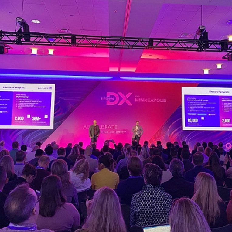 Presenters on stage at the Sitecore DX 2023 event in Minneapolis with audience and event graphics on screen.
