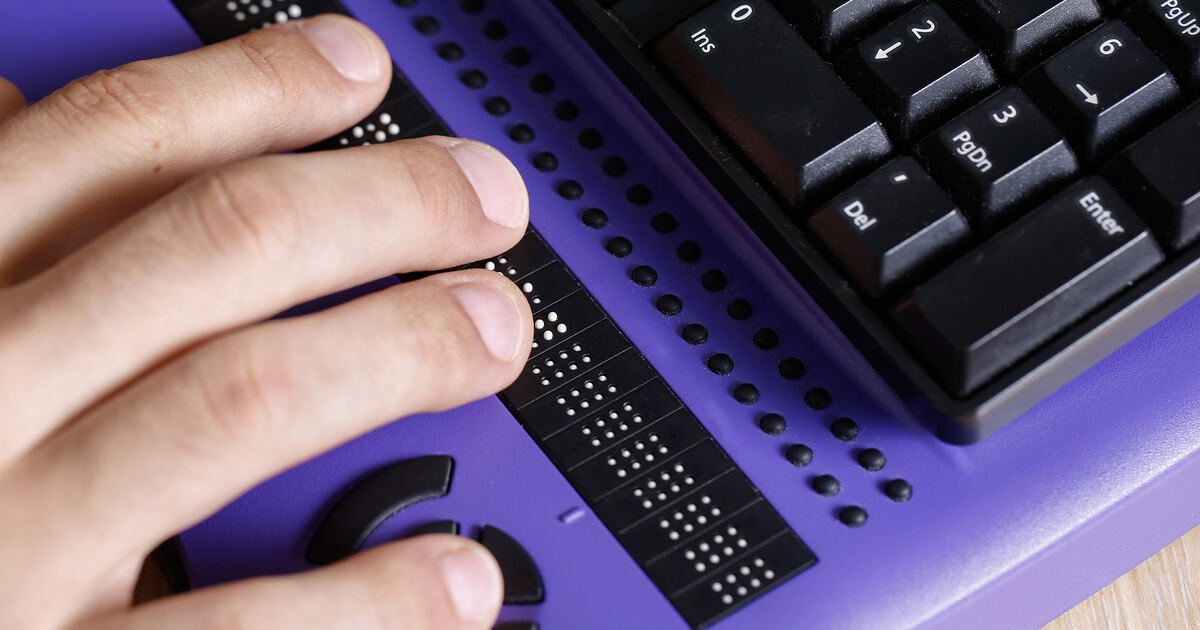 Hand reading a refreshable Braille display beside a keyboard, illustrating web accessibility and assistive technology use.