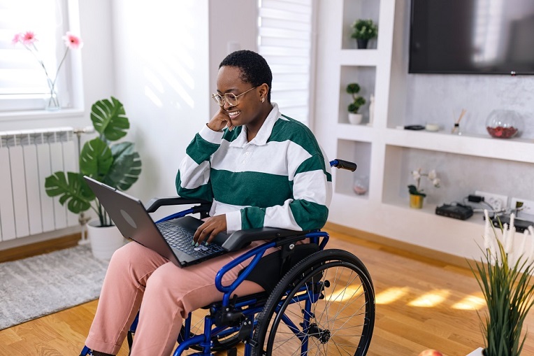 Smiling individual using a laptop while seated in a wheelchair in a bright, modern living room.