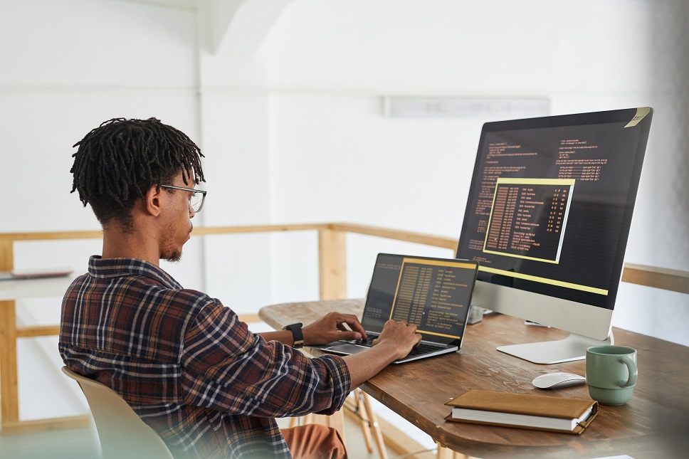 Programmer coding on a laptop and large screen monitor in a well-lit home office.
