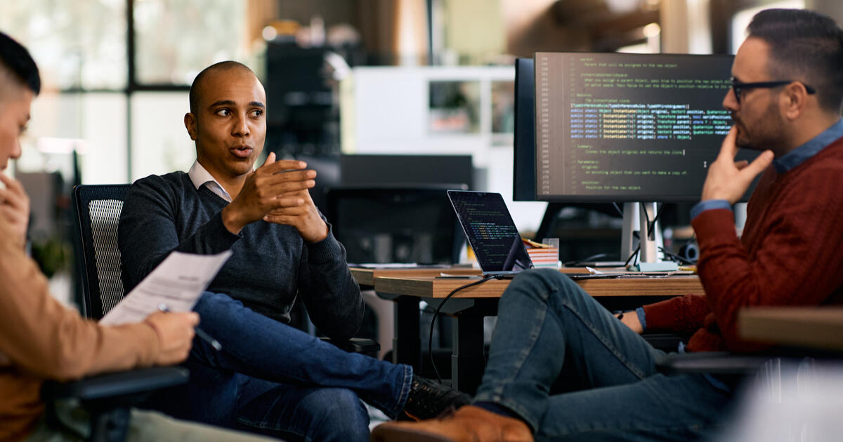 Software implementers meeting in an office with code displayed on monitors, illustrating collaboration and web development.