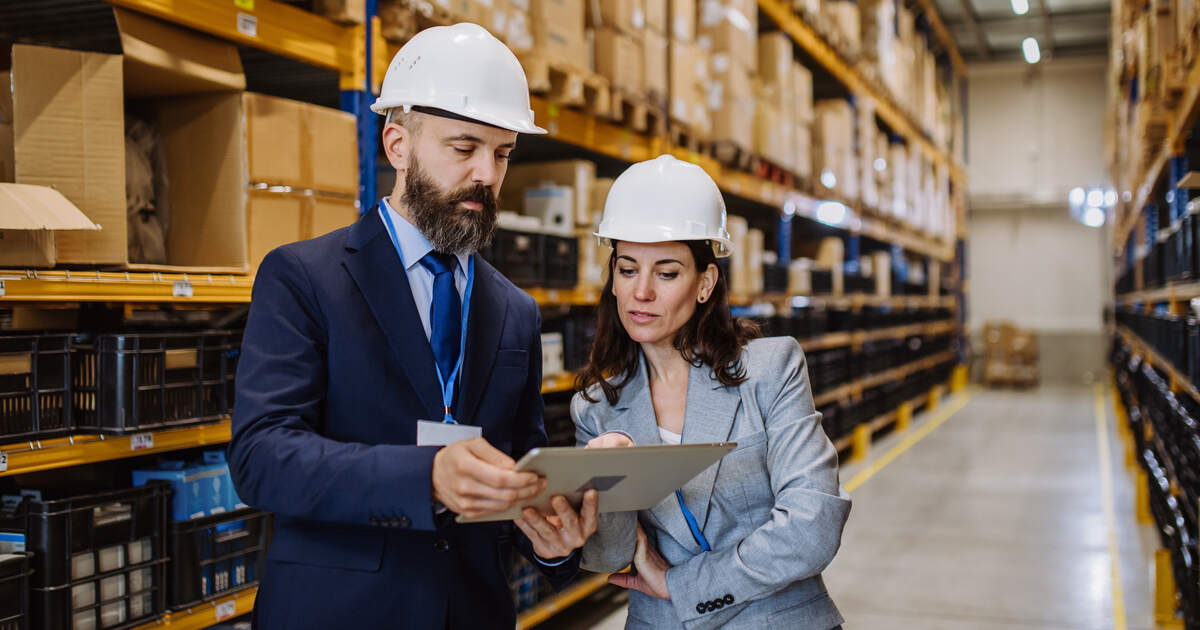 Marketers in hard hats reviewing warehouse inventory, representing wholesale distribution and digital marketing operations.