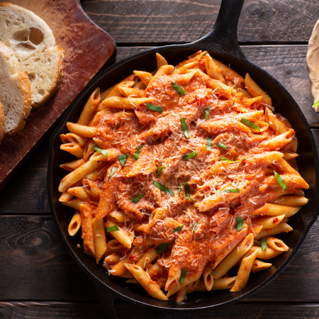 Skillet of baked penne pasta with tomato sauce and melted cheese, served with slices of bread on a wooden table.